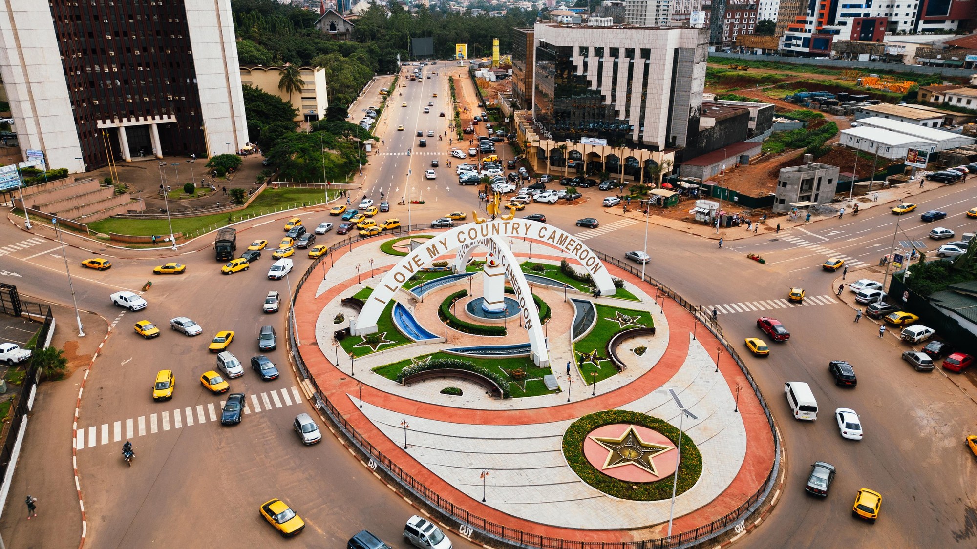 Aerial view of a busy circular roundabout with yellow taxis and palm trees in central Yaoundé