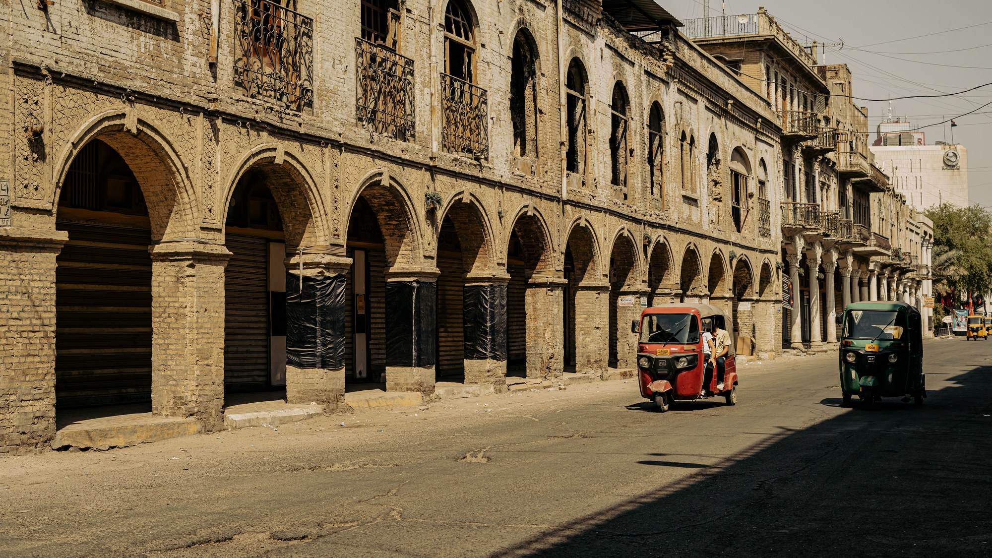 Bustling pedestrian street in historic Baghdad with architectural brick arches and yellow motorized tuk-tuks