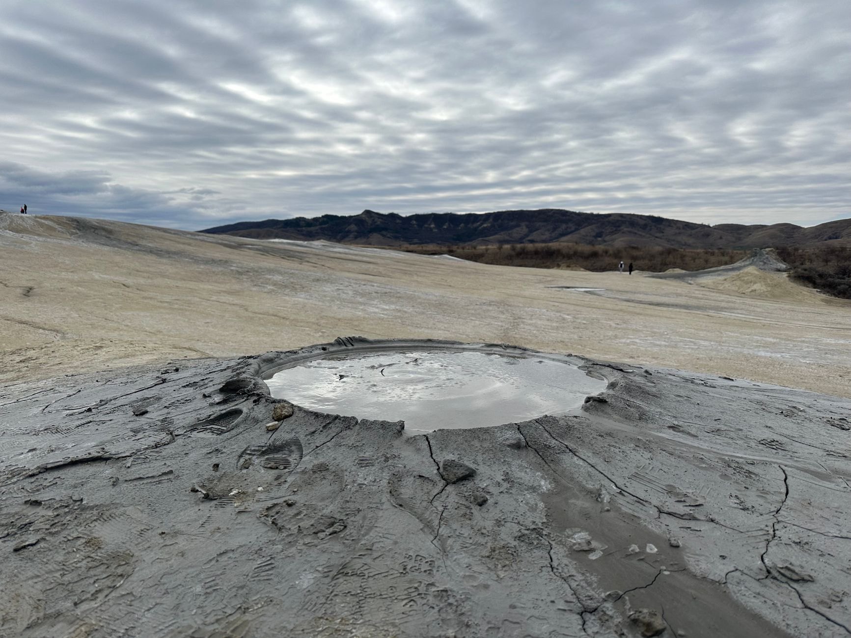 Small gray clay volcanic cone bubbling with cold liquid mud in a lunar desert landscape under wide skies