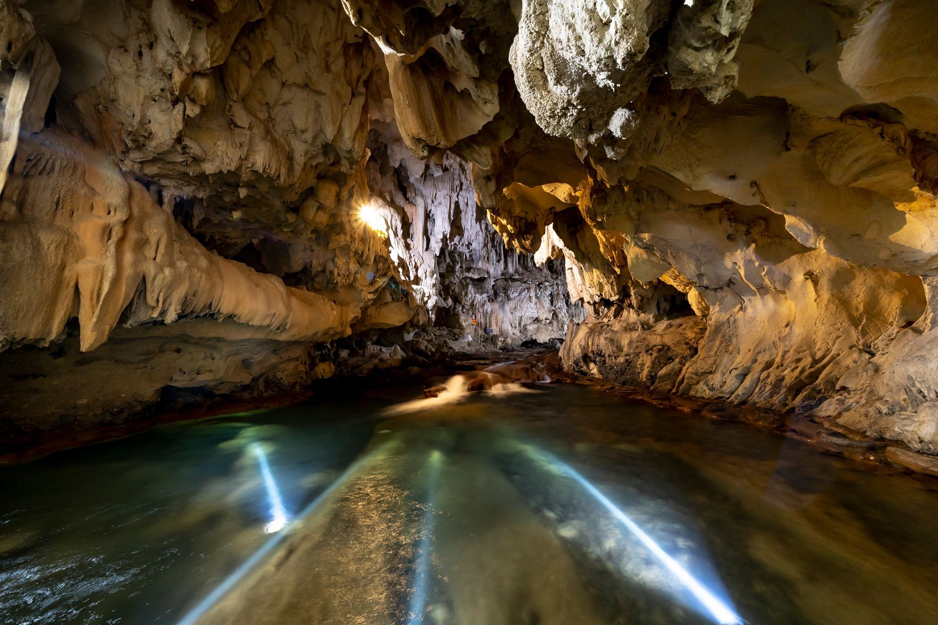 Massive cavernous limestone cave opening with clear river water flowing outward through dense tropical jungle foliage