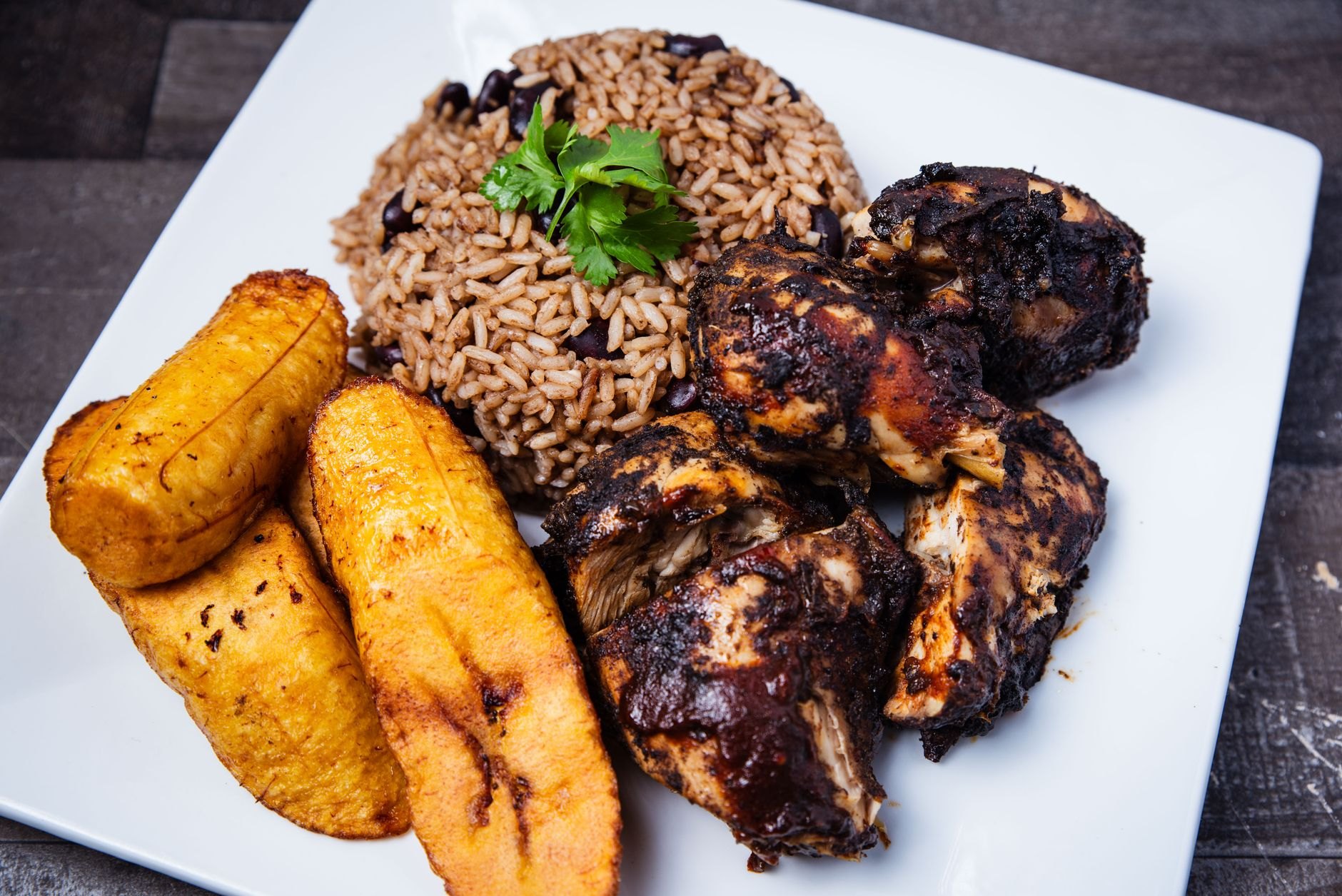 Generous plate of Belizean red recado stewed chicken served with coconut rice and beans, potato salad, and fried ripe plantains