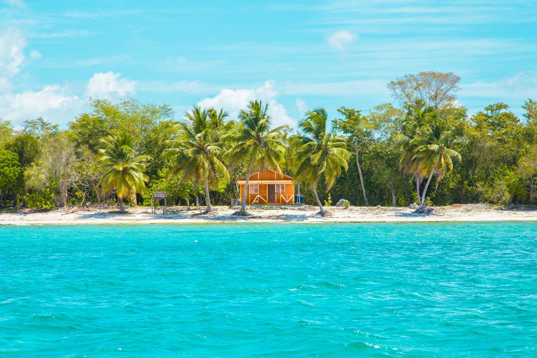 Brightly painted pastel wooden houses with wrap-around verandas lining a sandy unpaved street shaded by coconut palms