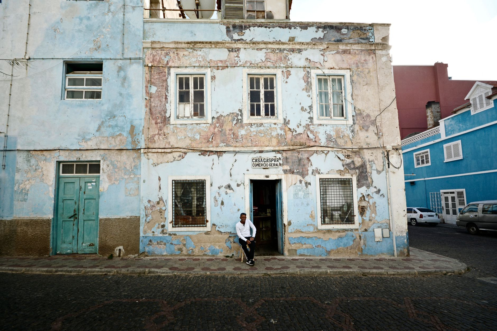 A man sitting outside a weathered pastel-colored colonial stone building in the cultural heart of Mindelo