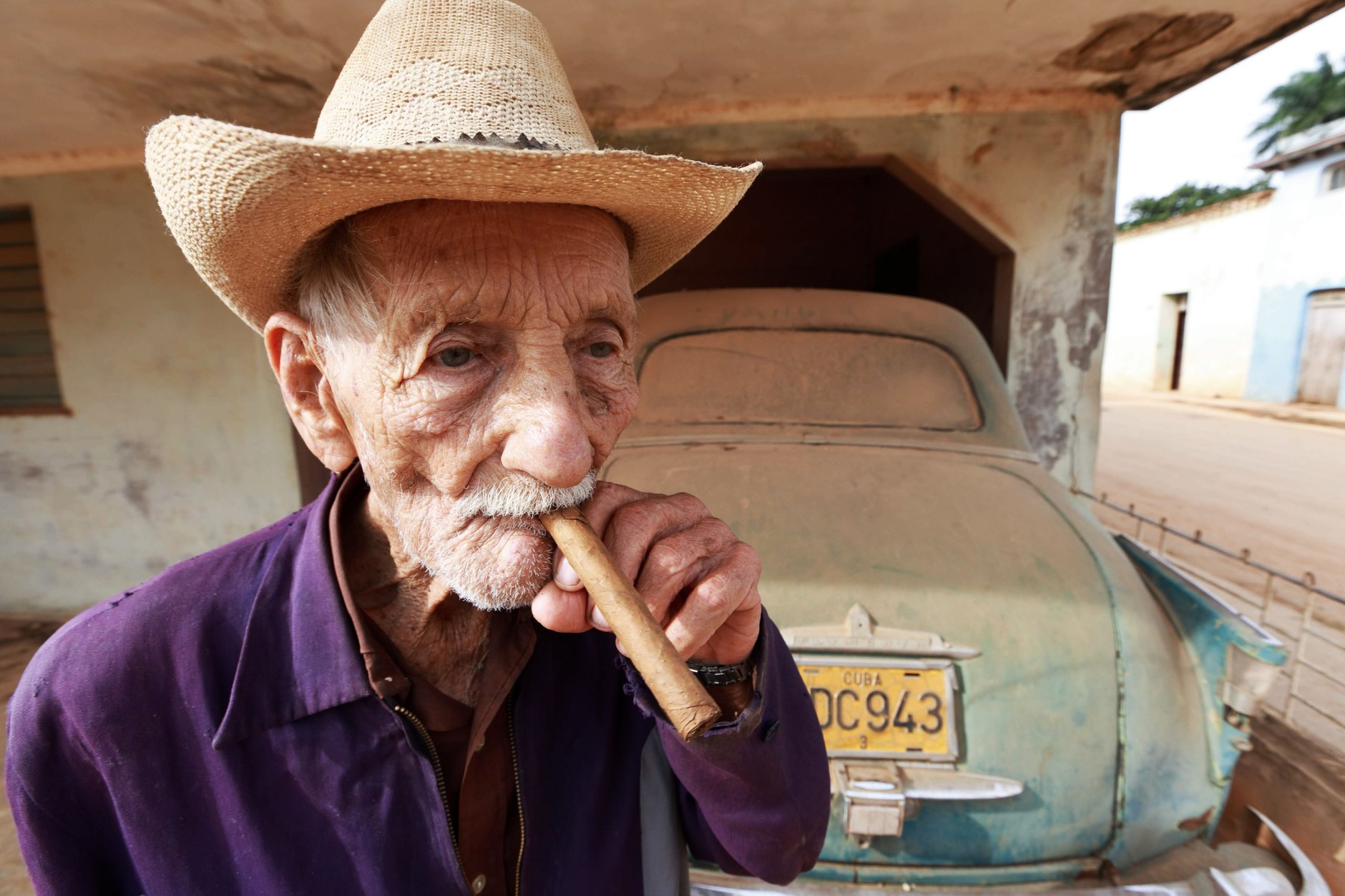 Senior Cuban resident in a straw hat beside an American vintage car on a city street in Cuba