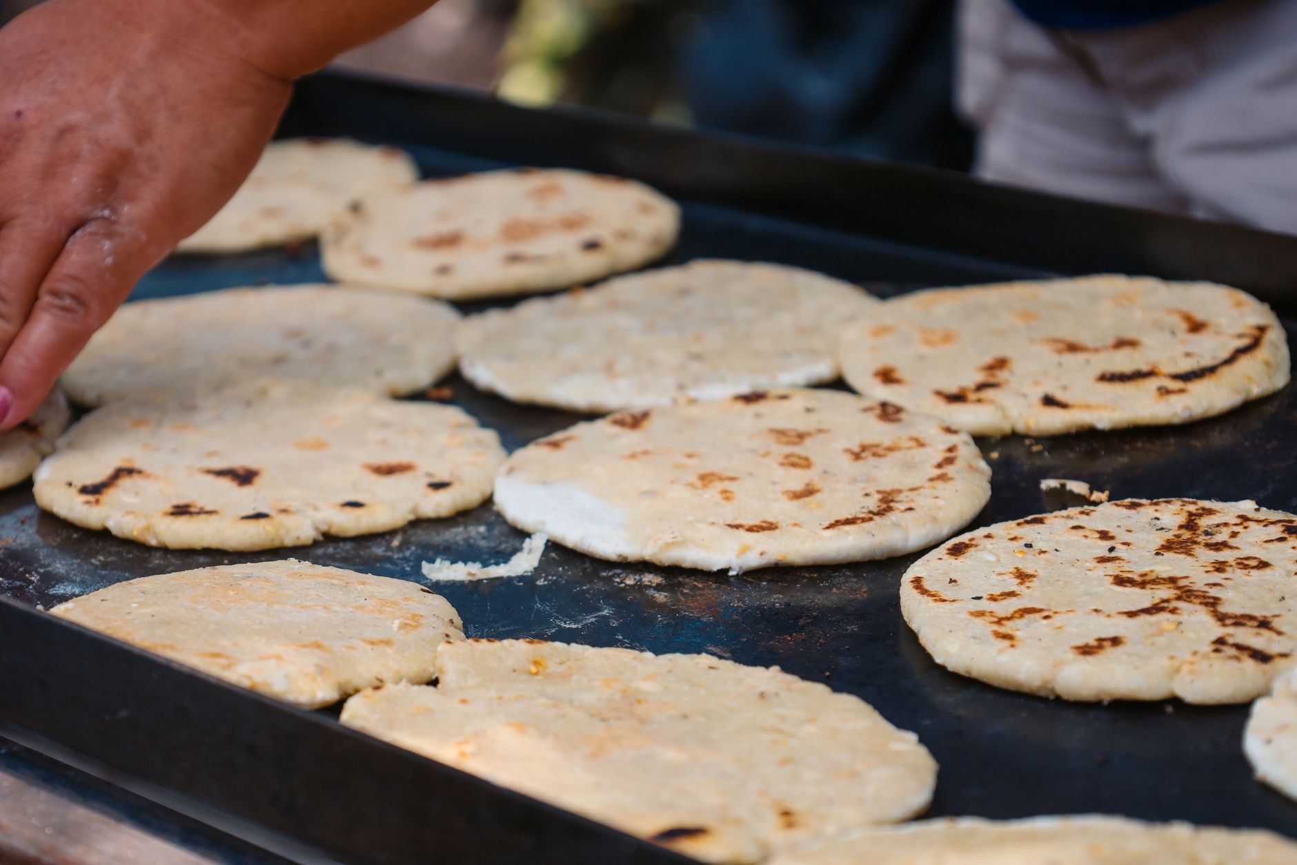 Stuffed corn pupusas sizzling on a round hot griddle alongside jars of fermented curtido cabbage slaw