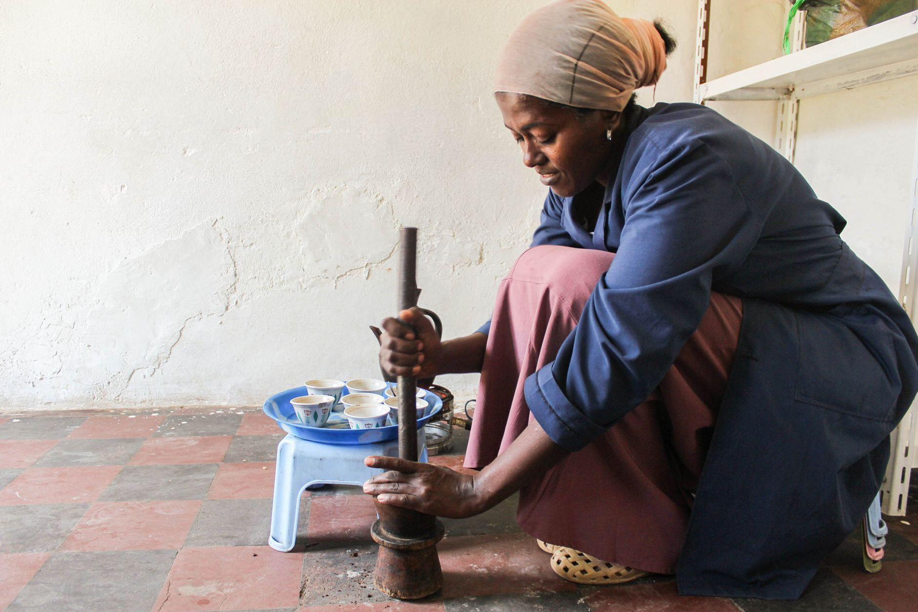 Woman in traditional attire grinding roasted coffee beans beside a clay jebena pot during a ceremonial brewing ritual