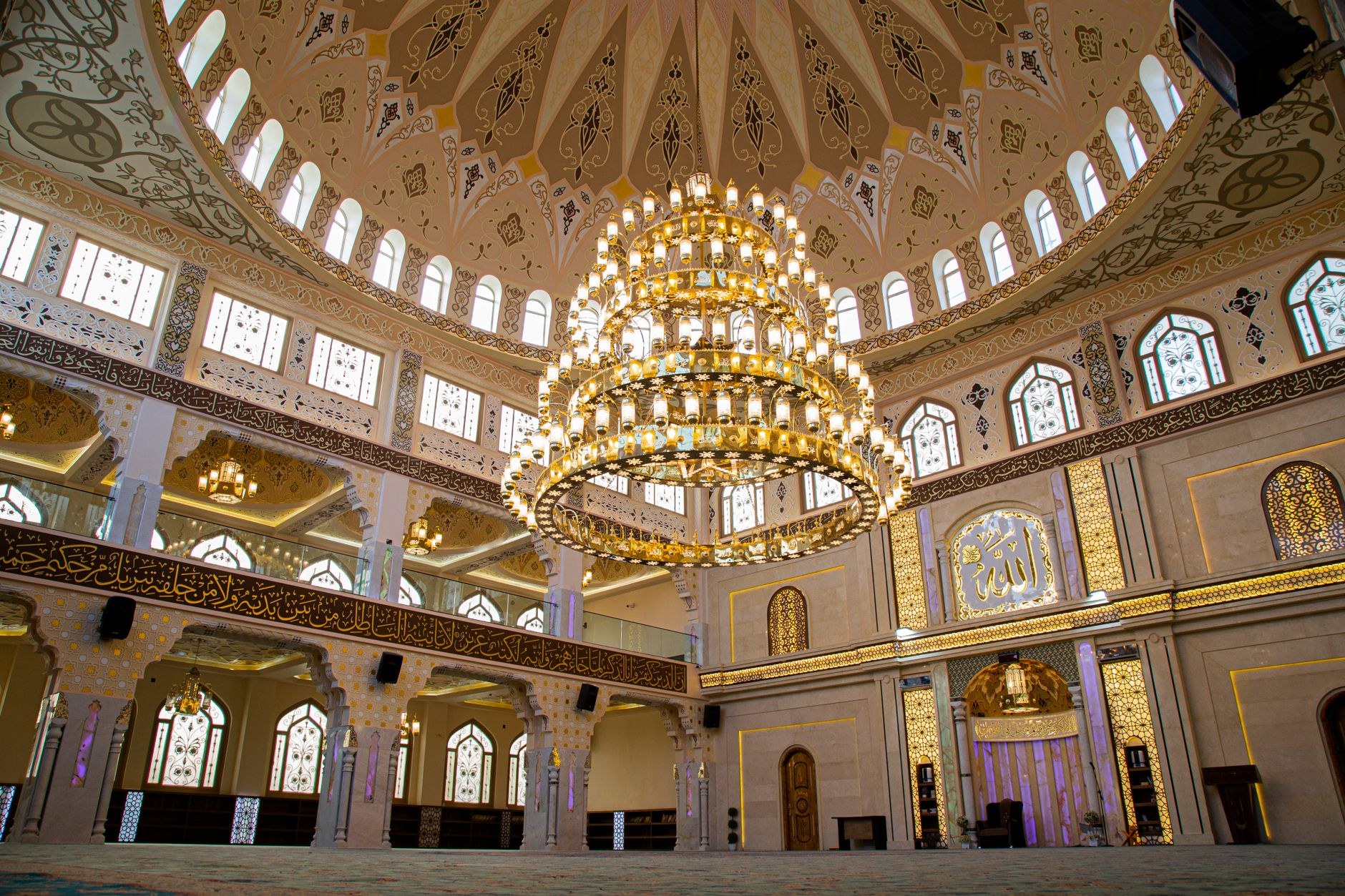 An expansive ornate mosque prayer hall with intricate blue and gold dome mosaic and grand chandelier