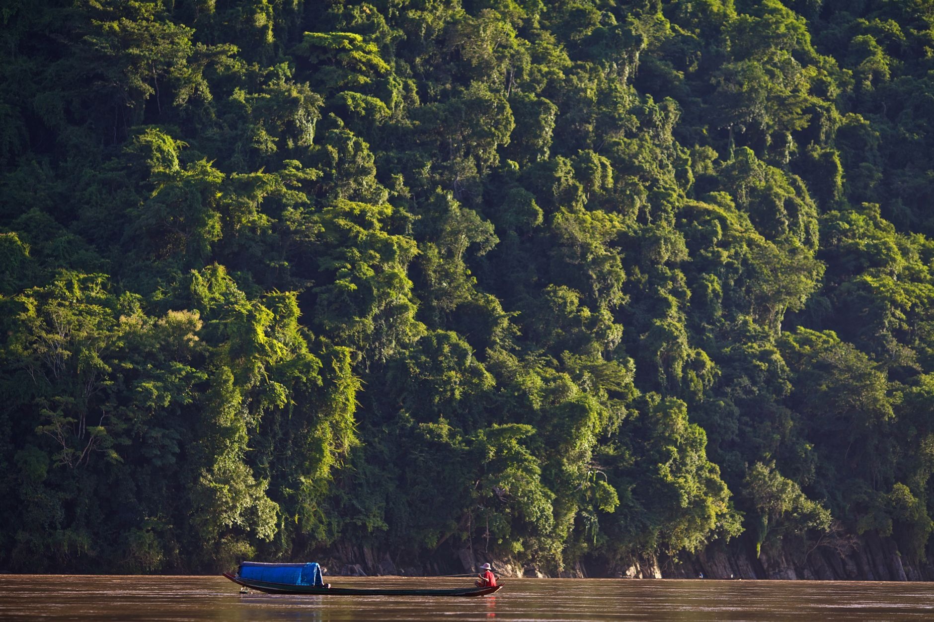 Traditional wooden passenger boat navigating the wide brown waters of the Mekong River past dramatic karst cliffs