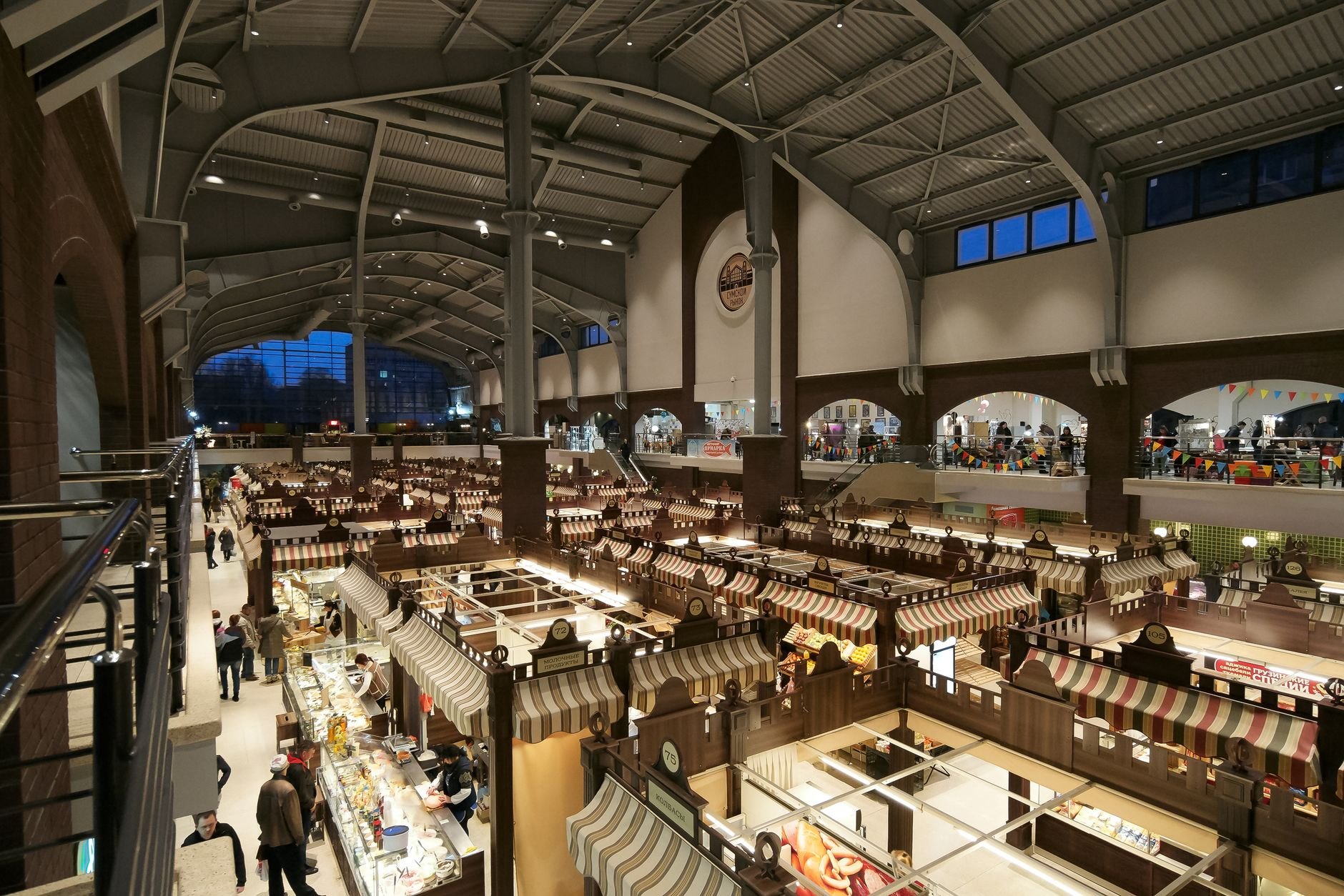 Spacious vaulted interior of a historic market pavilion with vendors displaying fresh fruits, pickles, and artisanal cheese