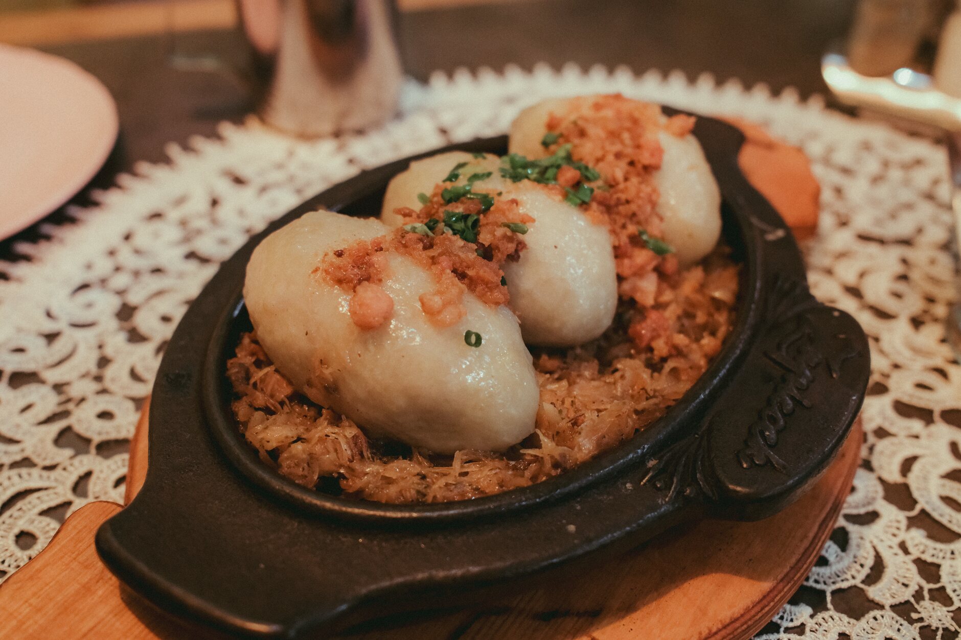 Oval potato dumplings topped with crisp bacon and herbs, served in a skillet on a lace tablecloth
