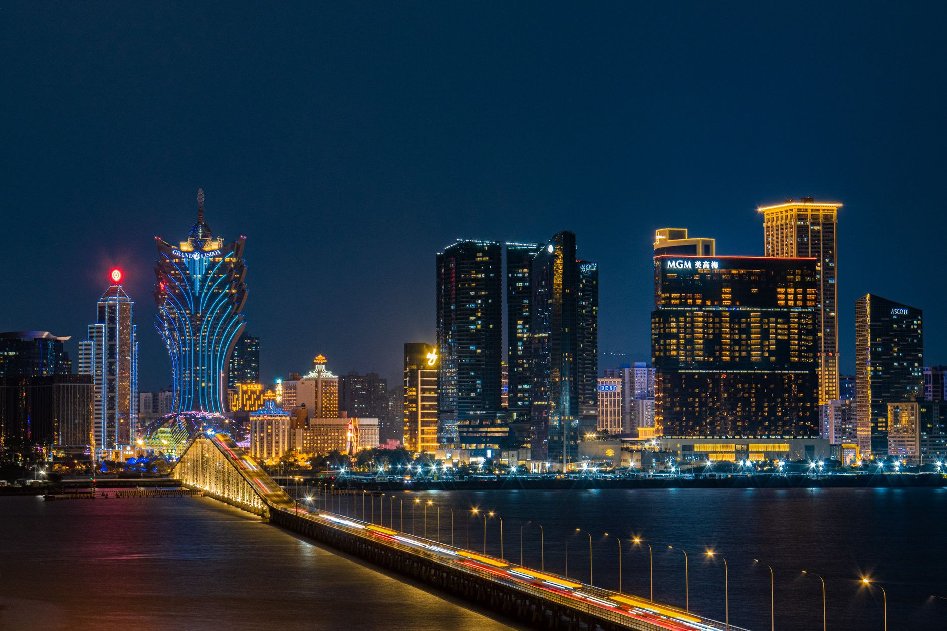 Panoramic nighttime view of Macao waterfront featuring illuminated bridge and glowing contemporary hotel towers reflecting across the harbor