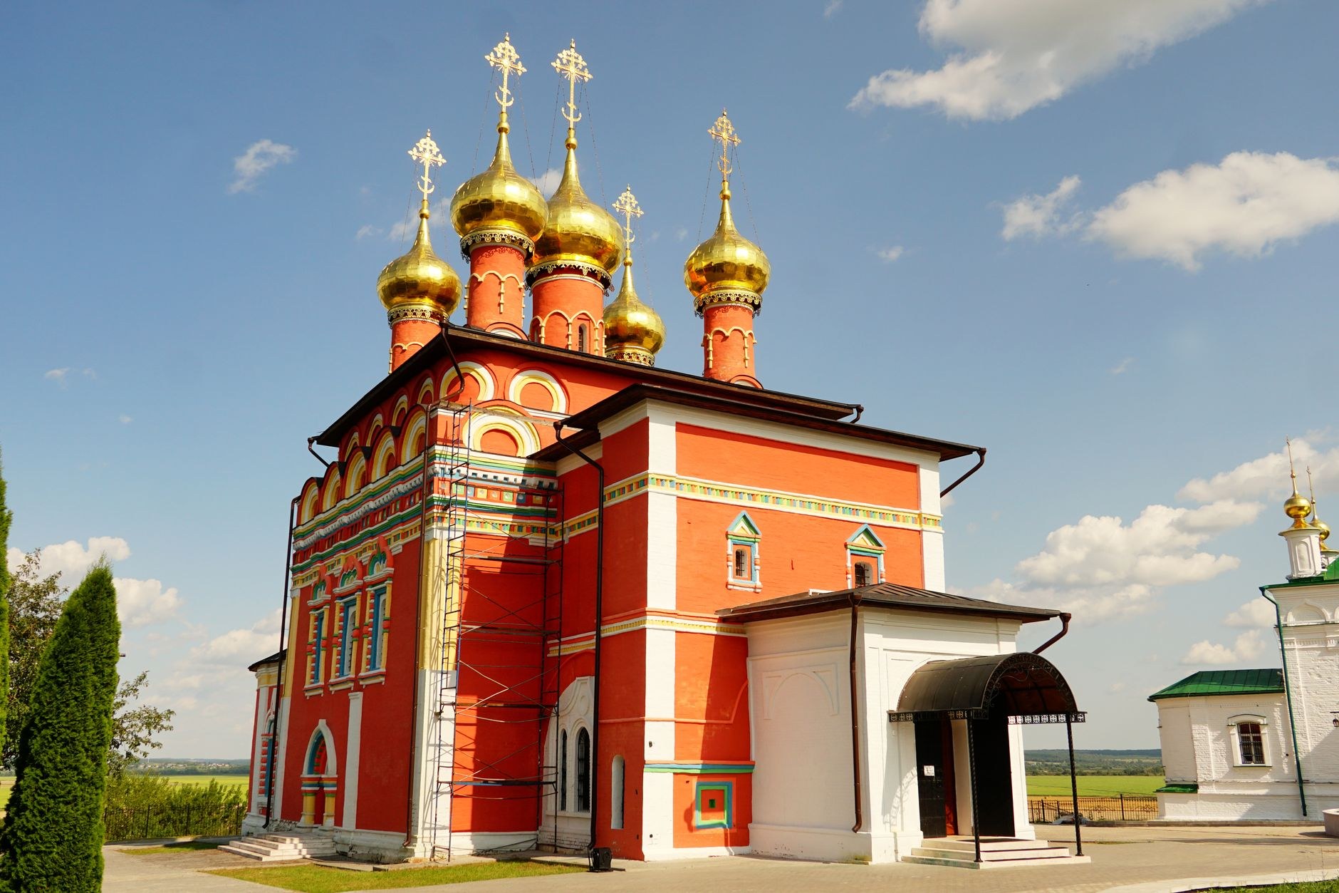An ornate baroque Orthodox monastery with crimson walls, white columns, and gilded onion domes in a manicured garden