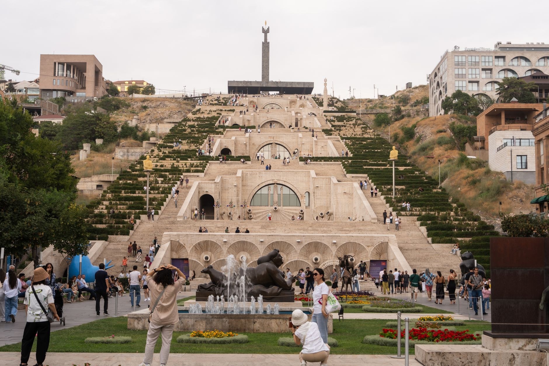 A monumental white stone cascade staircase with fountains descending through a lush park toward a peaceful lake