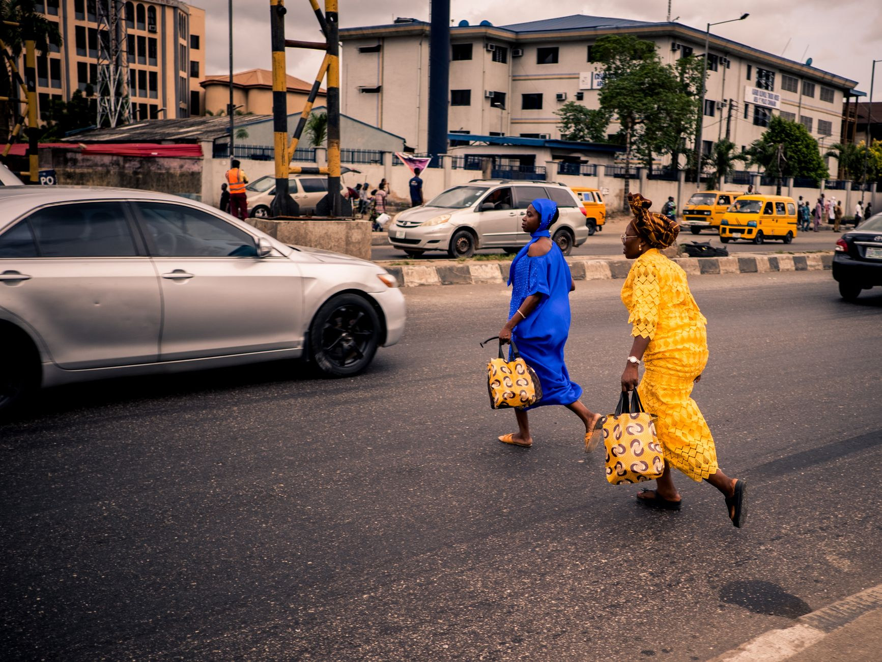 Two women wearing elegant traditional West African attire walking across a sunny urban street in Lagos