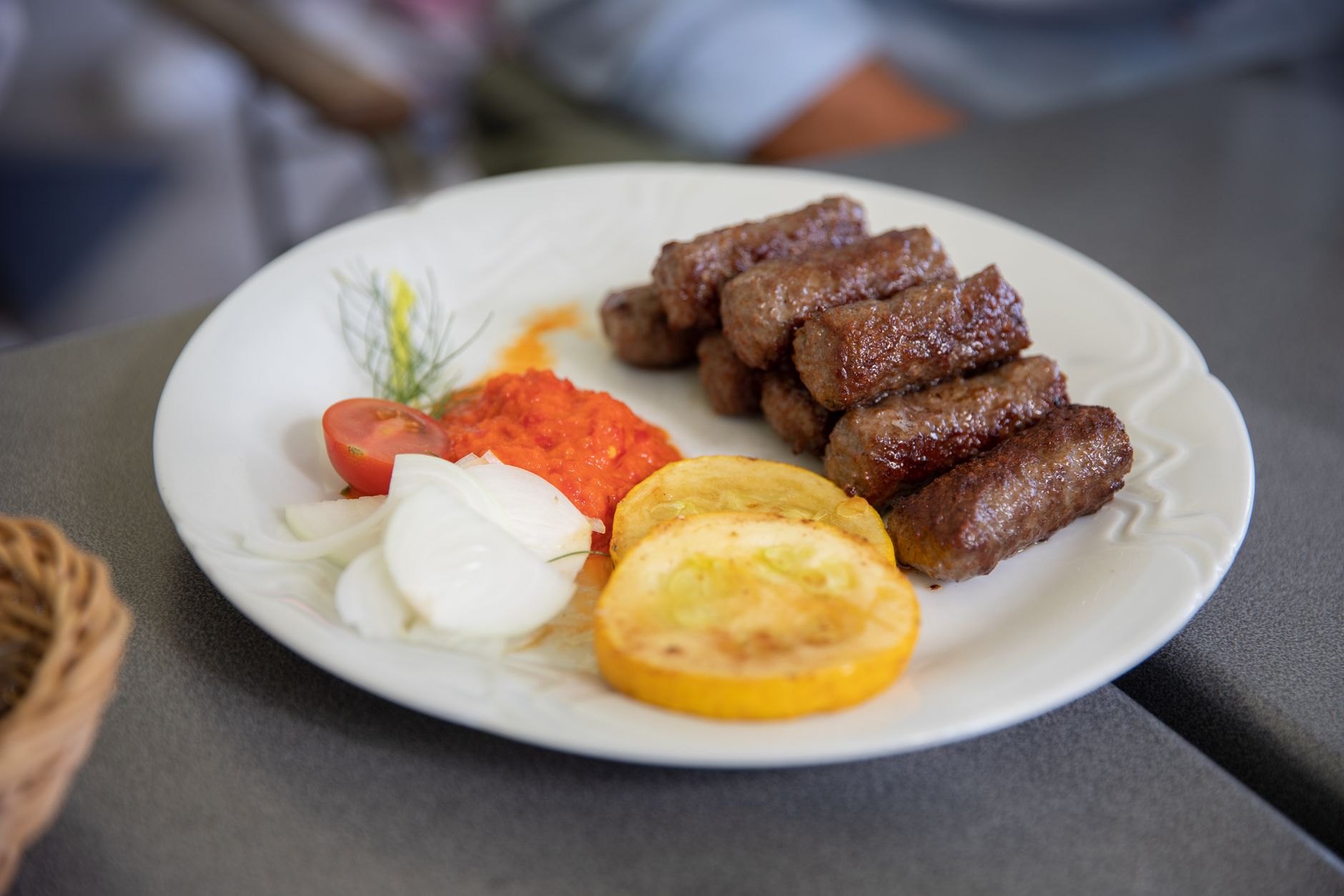 Platter of grilled Serbian ćevapi sausages served on warm somun flatbread with chopped raw onions, creamy kajmak, and red pepper ajvar