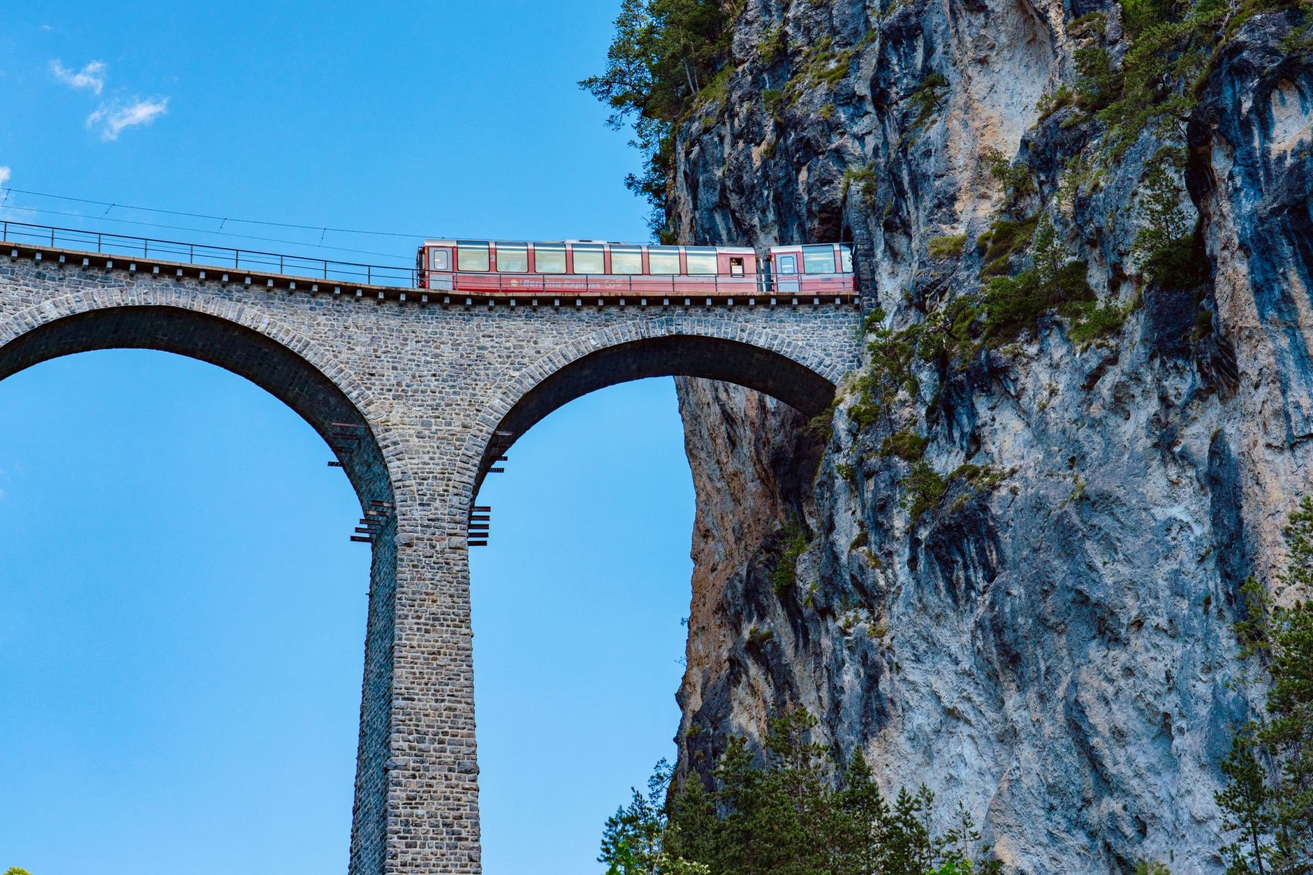 Red panoramic Swiss passenger train crossing a dramatic curved stone viaduct through alpine terrain