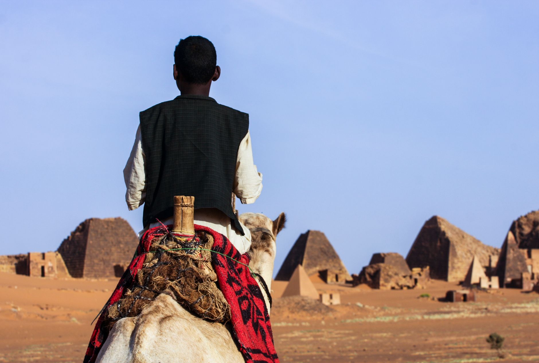 Bedouin rider leading a dromedary camel across desert sands toward ancient Kushite pyramids