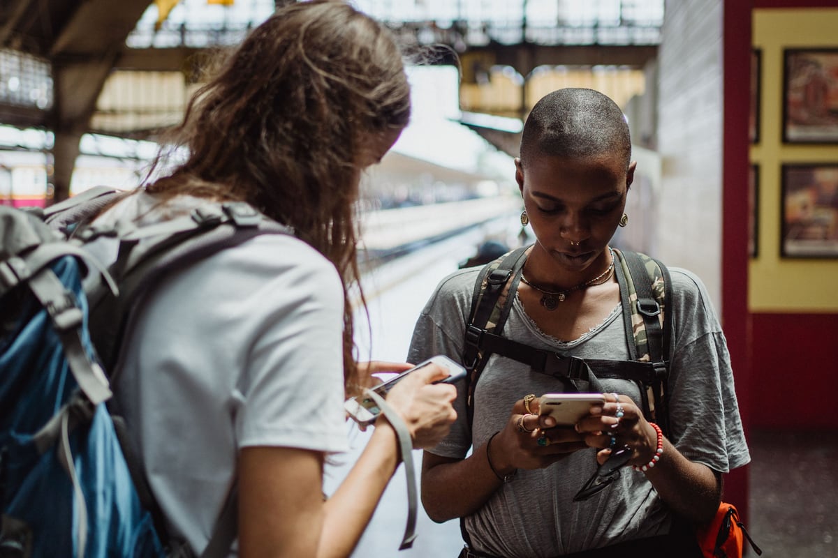 Two travelers with backpacks checking navigation and travel details on their smartphones at a train station platform.
