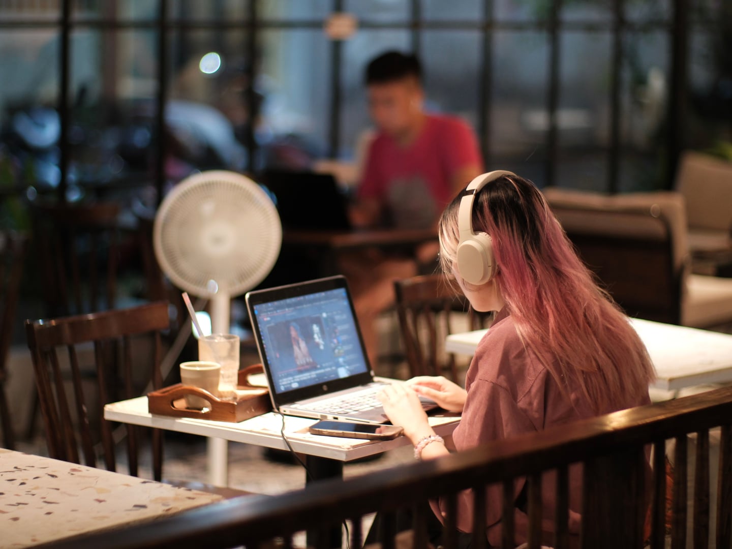 A creator wearing headphones editing travel video footage on a laptop in a café workspace.