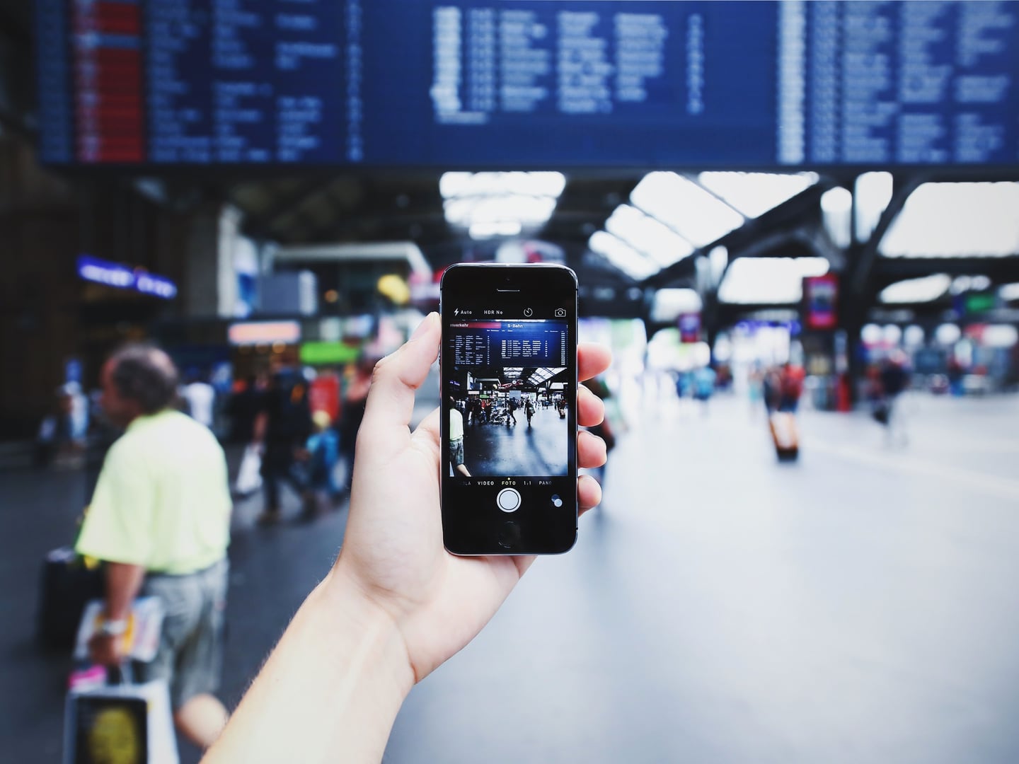 A traveler holding a smartphone filming a bustling transit station with travelers in the background.