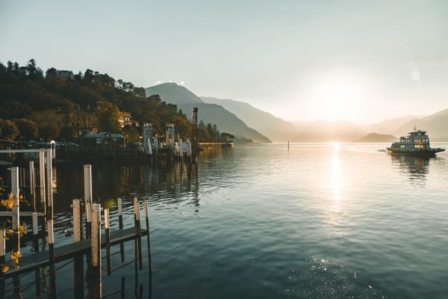 A passenger ferry glides toward the landing dock at golden hour on Lake Como in Varenna, Italy, framed by calm water and distant alpine peaks.
