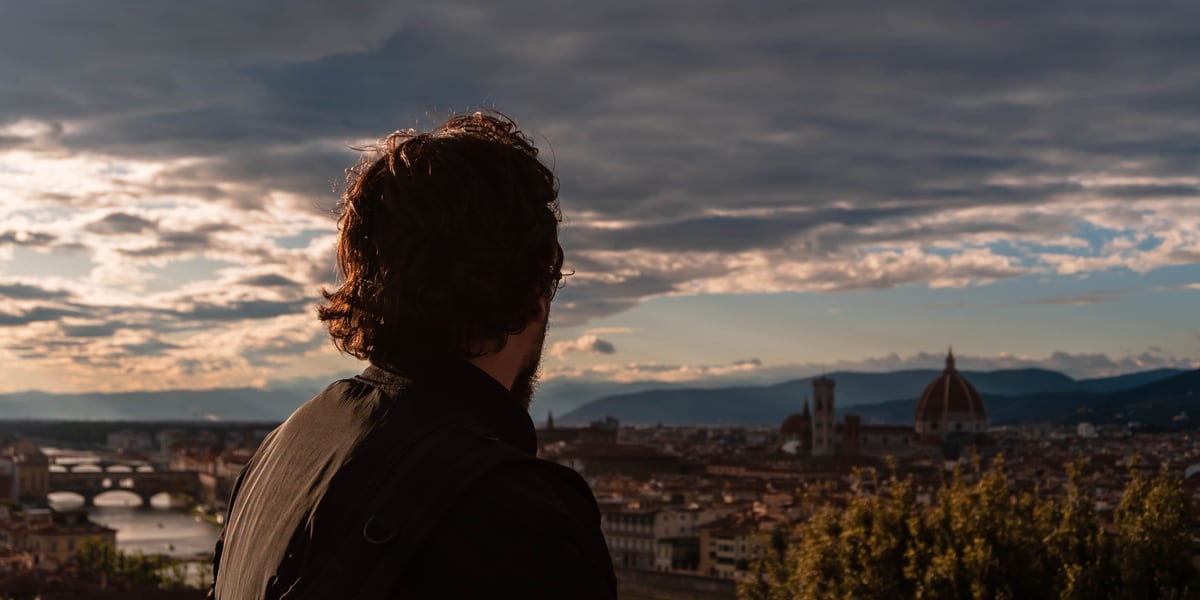 A traveller with a backpack looks out over a historic city skyline with warm sunset light on the clouds