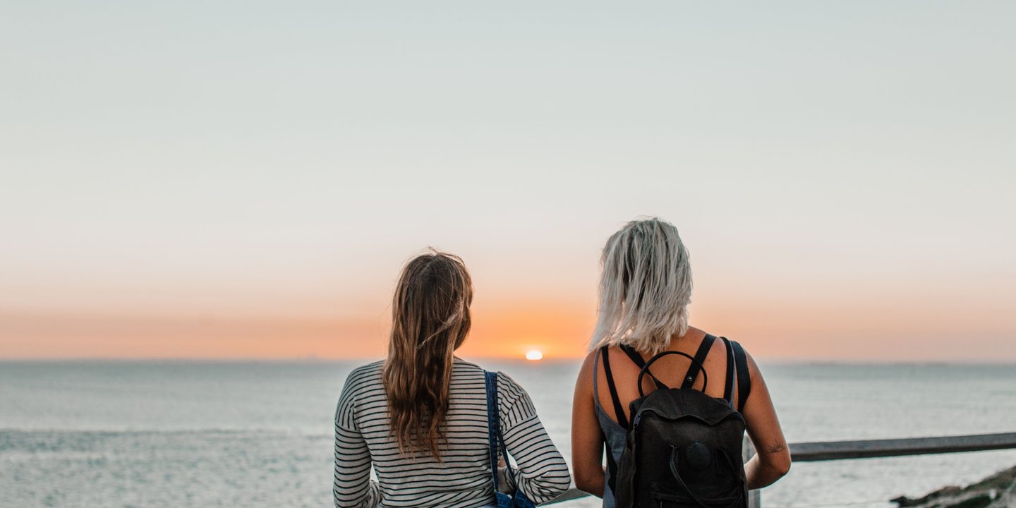 Two travelers with backpacks watching a serene ocean sunset from a coastal viewpoint