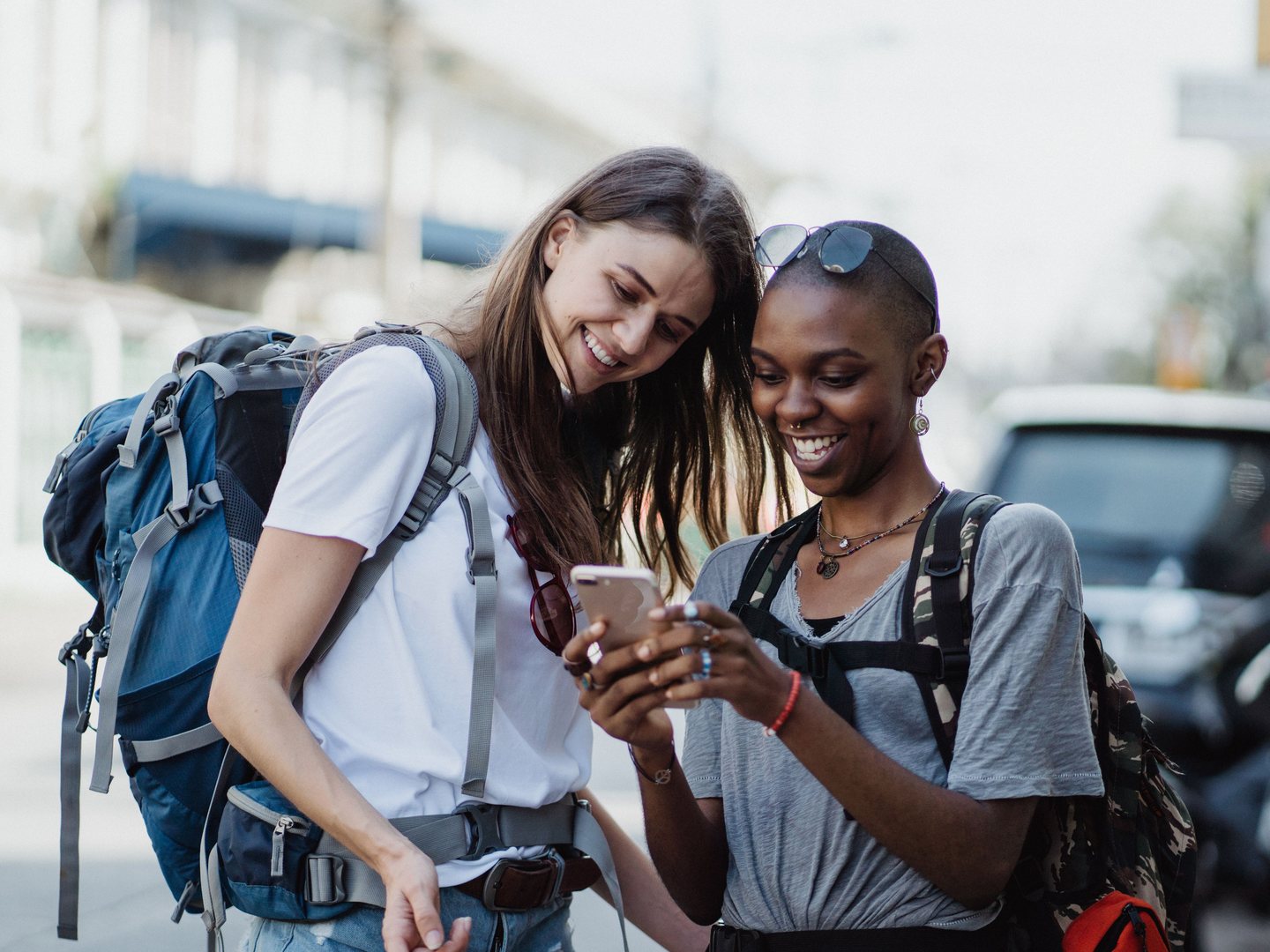 Two friends traveling with backpacks smiling and looking together at a smartphone on a city street