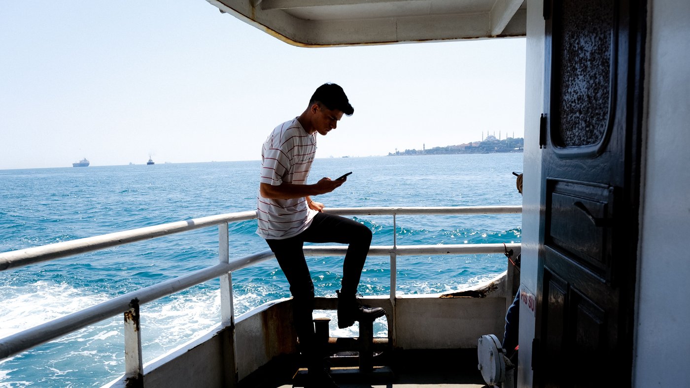 A passenger stands on an open boat deck looking at a smartphone, with sea behind him