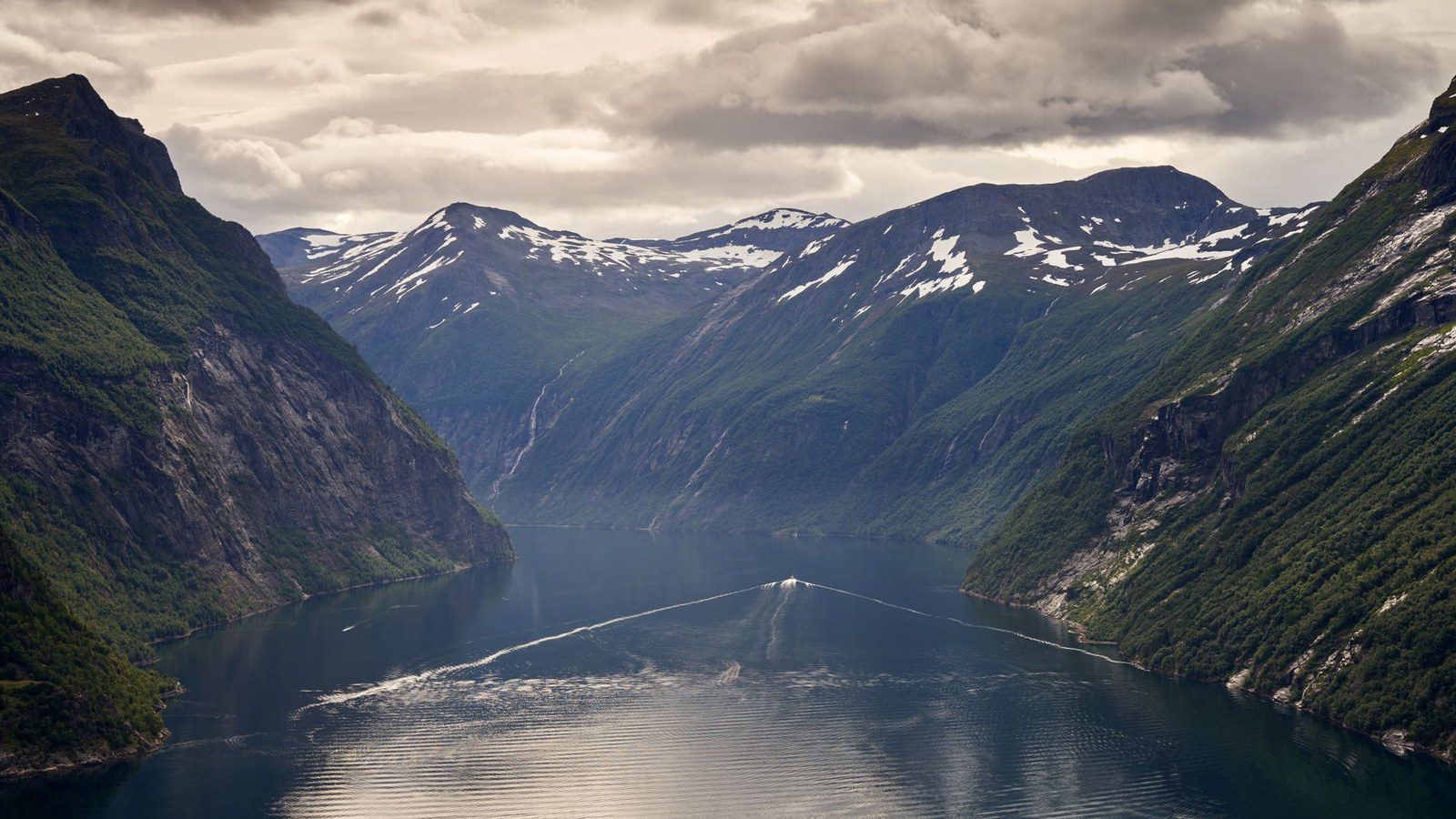 Breathtaking view of a Norwegian fjord with green mountains and snowy peaks under a cloudy sky.