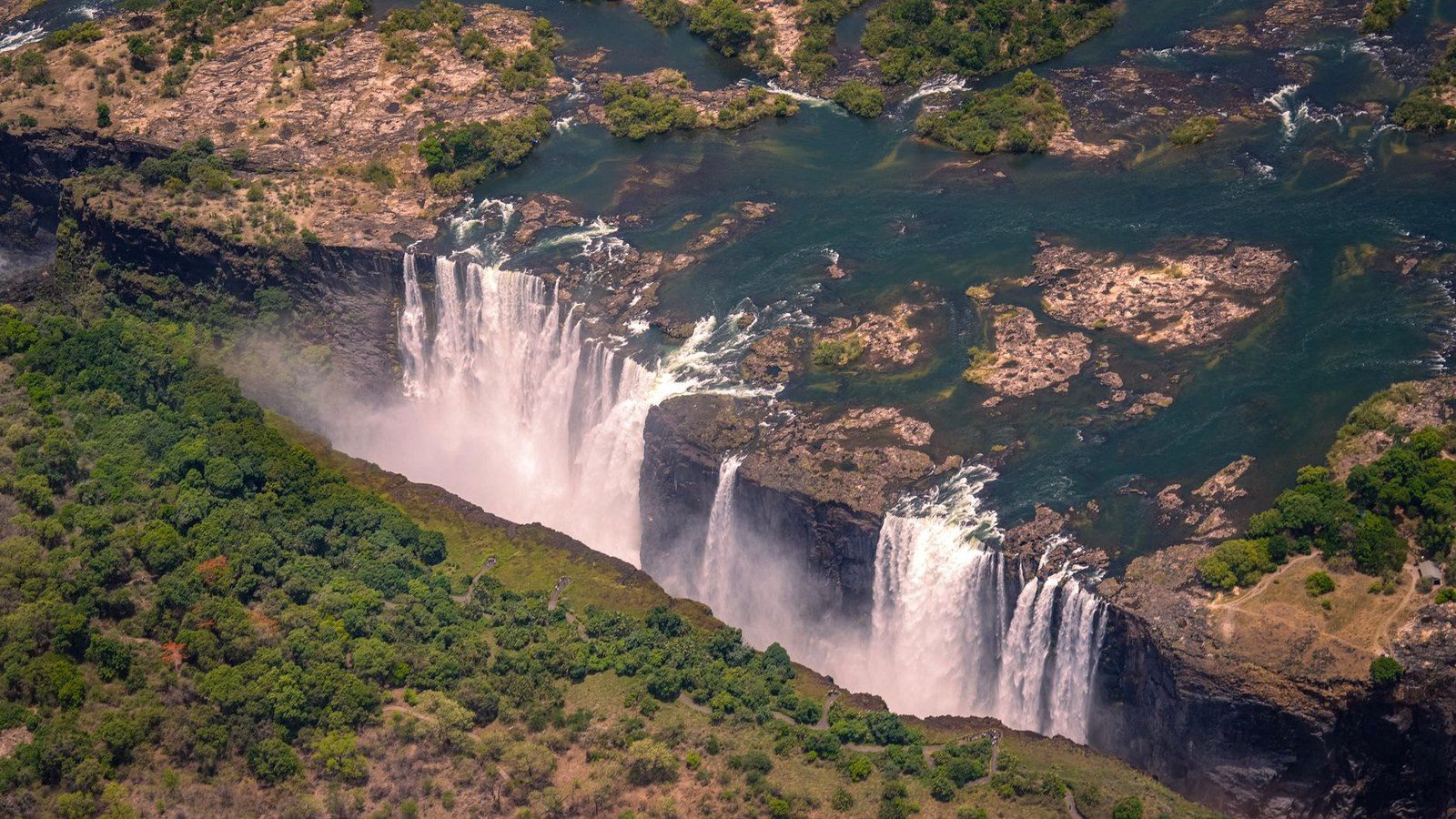 Dramatic curtain of water cascading into the deep basalt gorge of Victoria Falls with towering mist spray under blue sky